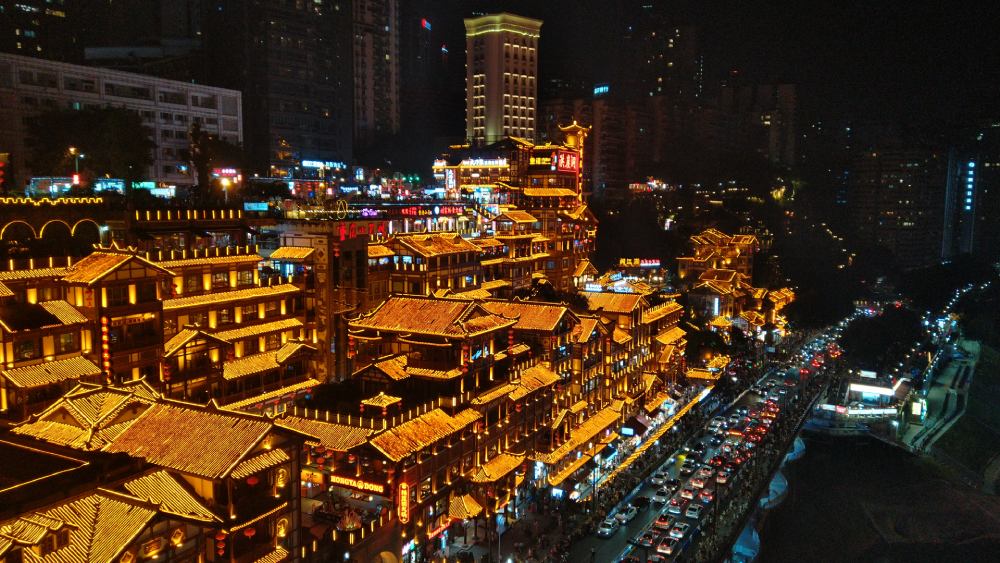 Complejo de edificios tradicionales iluminados con luces doradas en Hongya Cave, rodeado por la ciudad moderna de Chongqing durante la noche.